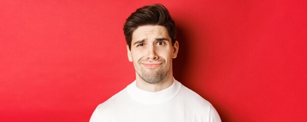 Close-up van een knappe man die zich ongemakkelijk voelt, grimassen trekt en naar iets onaangenaams kijkt, staande over een rode achtergrond