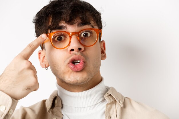 Close-up van een leuke en grappige kerel in een bril die naar brilmonturen wijst, een optiekwinkel aanbeveelt, staande op een witte achtergrond.