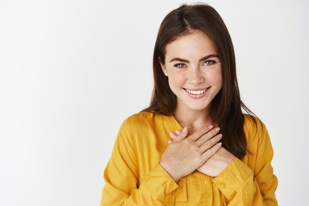 Close-up van een mooie vrouw die je bedankt, handen op het hart houdt en blij glimlacht, staande over een witte muur