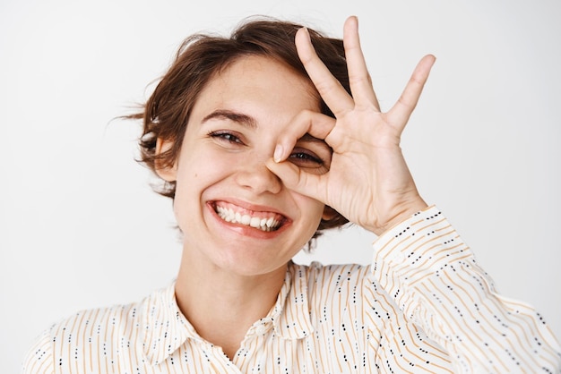 Close-up van een natuurlijke jonge vrouw die een goed teken op het oog laat zien en glimlachend iets goedkeurt dat een product aanbeveelt dat op een witte achtergrond staat