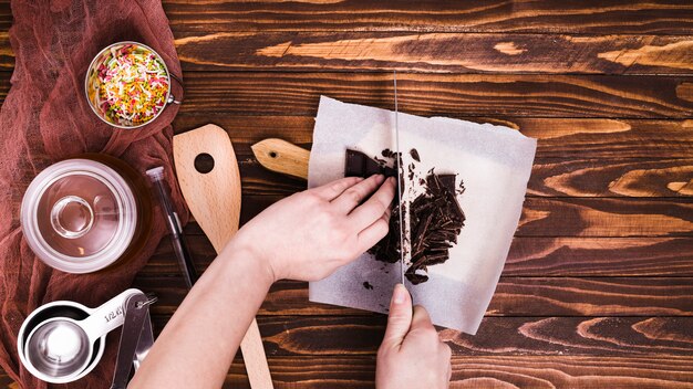 Close-up van een persoon die de chocoladereep met mes op papier over de houten lijst snijdt