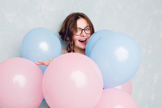 Close-up van een schattig brunette meisje dat in een studio staat, breed lacht en zich verstopt tussen blauwe en roze ballonnen. Ze draagt een zwarte bril en heeft gevlochten haar. Ze heeft plezier.