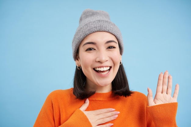 Close-up van een schattig Koreaans meisje met een muts steekt een hand op en stelt zichzelf voor, begroet je met een glimlach en