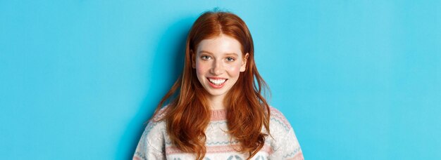 Close-up van een schattig roodharig meisje in een trui die blij glimlacht naar de camera die tegen een blauwe achtergrond staat