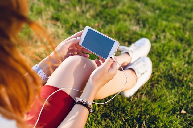 Close-up van een smartphone in handen van het meisje. Meisje zit op het groene glas, gekleed in een rode rok en witte sneakers