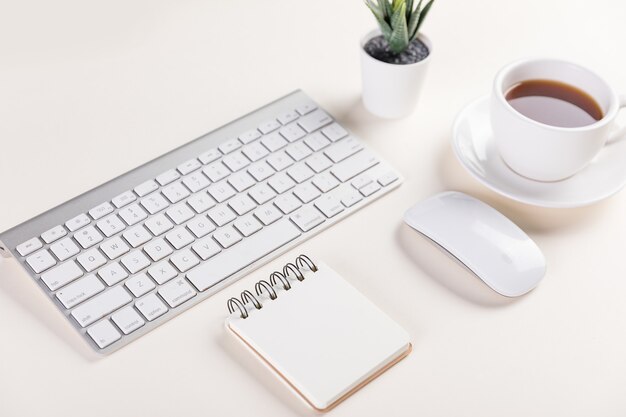 Close-up van een toetsenbord, notitieblok, computermuis, een kop warme koffie en een plant op witte tafel