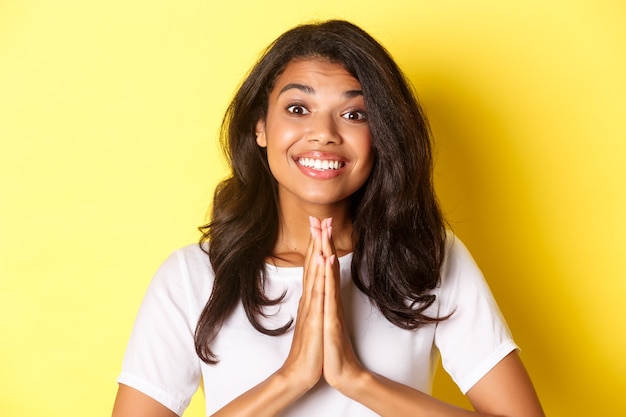 Close-up van een vrolijk afro-amerikaans meisje, glimlachend en dank je wel, druk de handen op de borst in een gebedsgebaar, staande over gele achtergrond