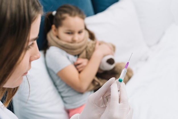 Close-up van een vrouwelijke arts handen vullen de spuit met medicijnen met wazig meisje zittend op bed