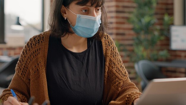 Close-up van een werknemer die met een laptop werkt en aantekeningen maakt op papieren met grafieken, met een gezichtsmasker. Vrouw die computer gebruikt om aan een zakelijk project te werken tijdens de pandemie van het coronavirus.