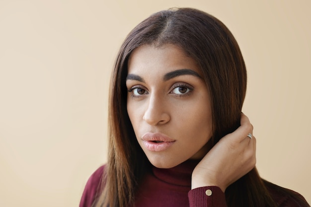 Close-up van geweldige jonge donkere vrouw met lang donkerbruin haar en schone perfecte huid poseren, kijken met lippen lichtjes gescheiden, haar kapsel aanpassen. Horizontaal schot