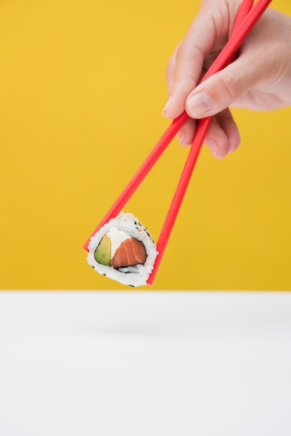 Close-up van iemands hand met sushi roll met rode stokjes tegen gele achtergrond