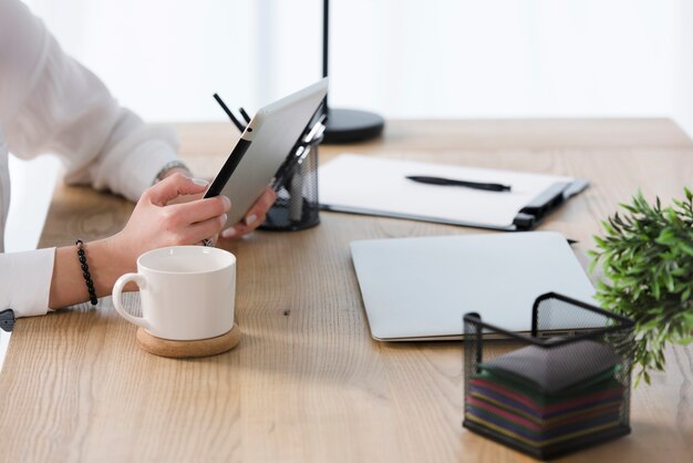 Close-up van jonge onderneemster die digitale tablet met koffiekop gebruikt; laptop op houten tafel