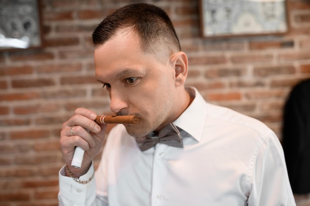 Gratis foto close-up van knappe man in stijlvol wit overhemd en vlinderdas die cubaanse sigaar vasthoudt en ruikt tijdens de huwelijksaangelegenheid