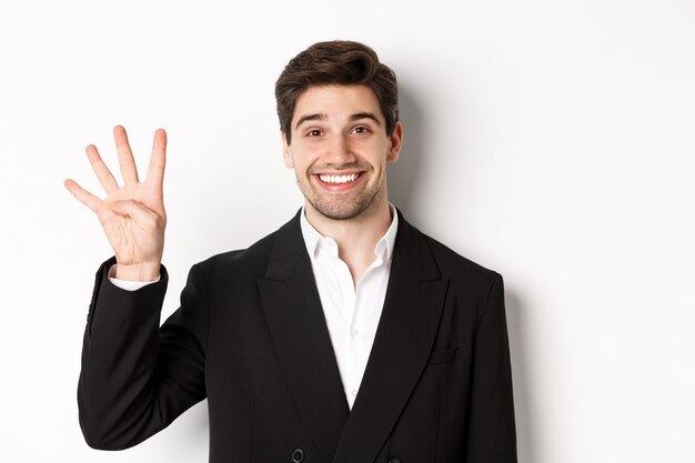 Close-up van knappe zakenman in zwart pak, verbaasd glimlachend, nummer vier tonend, staande op een witte achtergrond