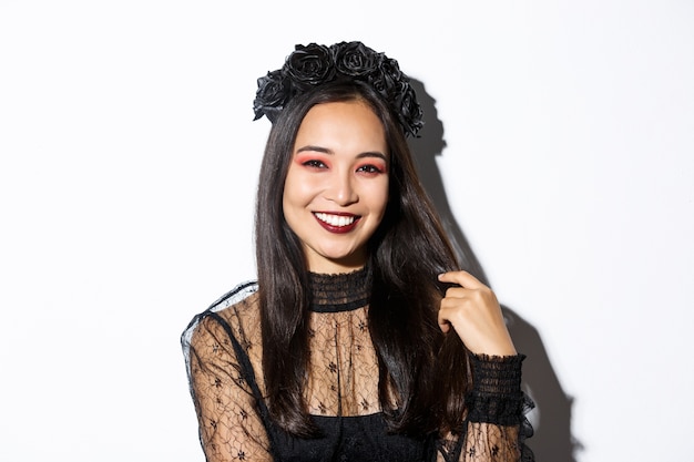 Close-up van mooie elegante aziatische vrouw in zwarte krans en gotische kanten jurk glimlachen, staande op witte achtergrond, gekleed voor halloween-feest.
