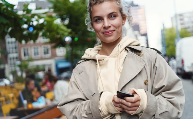 Close-up van mooie jonge vrouw die zich op straat met mobiele telefoon bevindt en gelukkig glimlacht.