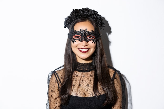Close-up van mysterieuze vrouw in gotische krans en zwart masker glimlachend in de camera, halloween vieren, staande op witte achtergrond.