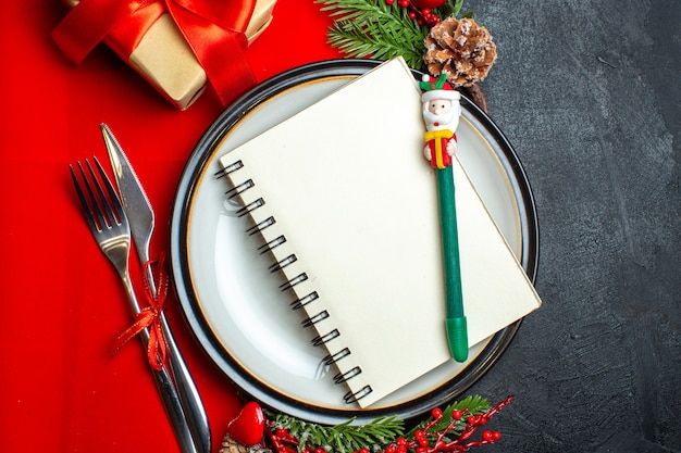 Close-up van Nieuwjaar achtergrond met spiraal notebook op diner plaat bestek set decoratie accessoires fir takken naast een geschenk op een rode servet op een donkere tafel