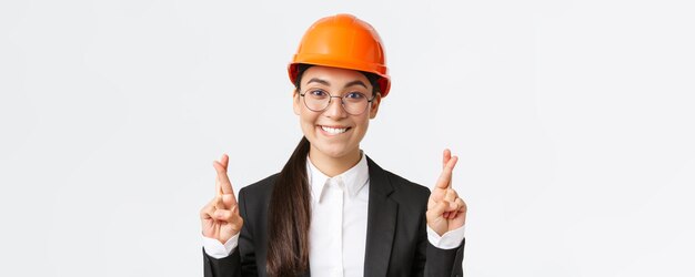 Close-up van opgewonden hoopvolle aziatische vrouwelijke ingenieur bouwmanager in veiligheidshelm en pak smekend voor relish kruis vingers veel geluk en bijtende lip verleidelijke witte achtergrond