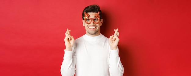 Close-up van opgewonden knappe man, met feestbril en witte trui, vingers kruisend voor geluk, kerst vieren en wachtende wens, in afwachting van geschenken, staande op rode achtergrond