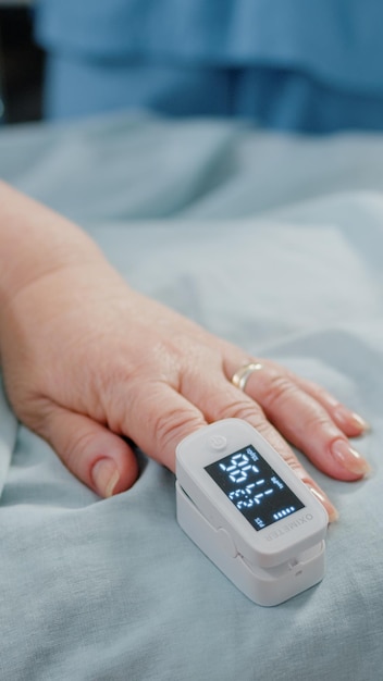 Close up van oximeter aan kant van senior patiënt tot in bed. Leeftijd persoon met elektronisch apparaat voor zuurstofverzadiging en polsdrukmeting op vinger. Volwassene met cardiologietool