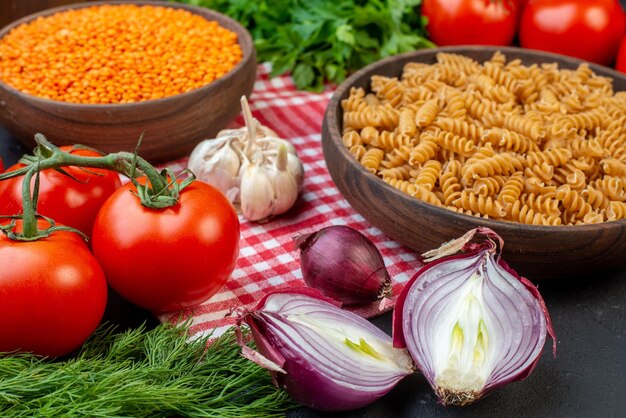 Close-up van rauwe pasta's op een houten snijplank op rode gestripte handdoek en verse groenten groen op donkere achtergrond