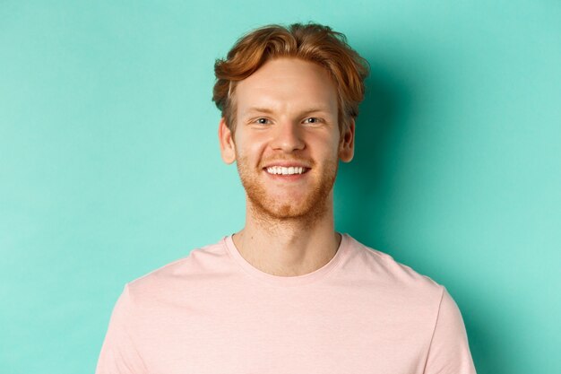 Close up van roodharige bebaarde man in roze t-shirt, glimlachend met witte perfecte tanden en camera kijken, staande op turkooizen achtergrond.