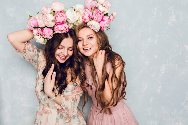 Close-up van twee mooie meisjes staan in een studio, die gek spelen met cirkels van bloemen op hun hoofd. Ze dragen lichte zijden jurken. Een is blond en een is brunette. Beiden hebben krullend haar.