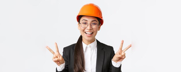 Close-up van vrolijke succesvolle vrouwelijke Aziatische ingenieur bouw architect in veiligheidshelm en pak met vredestekens en lachende kawaii staande witte achtergrond