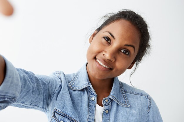 Close-up zeer gedetailleerd portret van charmante jonge Afro-Amerikaanse vrouw met zwart haar en een perfecte gezonde donkere huid, gekleed in een lichtblauw spijkerblouse, met een mooie schattige glimlach.