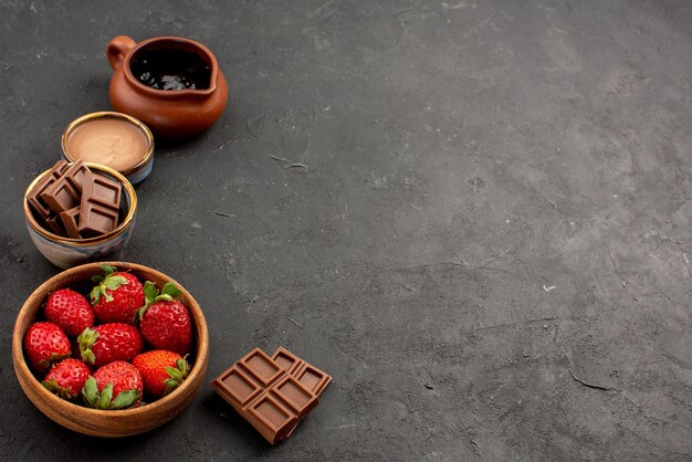 Close-up zijaanzicht aardbeien, aardbeien en chocoladeroom in kommen en chocoladerepen aan de linkerkant van de donkere tafel