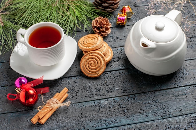 Close-up zijaanzicht een kopje thee koekjes naast het kopje thee kaneelstokjes een theepot vuren boom met kegels en kerstspeelgoed op de houten tafel