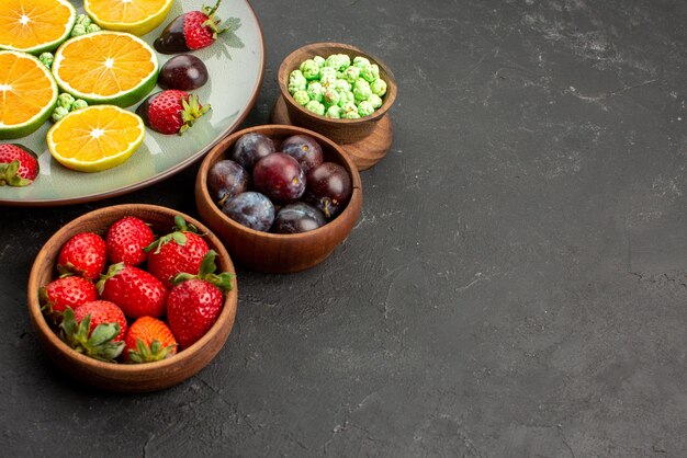 Close-up zijaanzicht fruit met chocolade bedekte aardbei gehakte oranje en groene snoepjes naast kommen met verschillende soorten fruit en snoep aan de linkerkant van de donkere tafel
