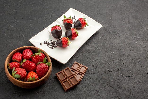 Close-up zijaanzicht met chocolade omhulde aardbeien kom met aardbeien en repen chocolade naast het bord met met chocolade omhulde aardbeien op de donkere tafel
