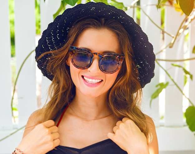 Close-up zonnige levensstijl portret van vrij lachende vrouw poseren in de buurt van witte muur, net afkomstig van het strand, stijlvolle zwarte hoed en elegante vintage zonnebril dragen.