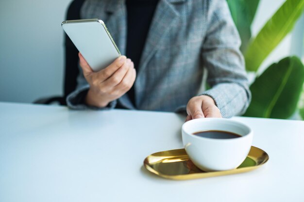 Close-upbeeld van een zakenvrouw die een mobiele telefoon vasthoudt en gebruikt terwijl ze koffie drinkt op kantoor