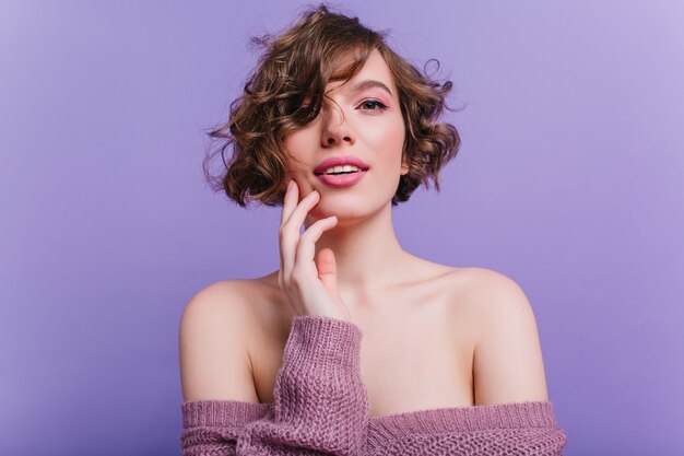 Close-upfoto van schattig vrouwelijk model met roze make-up. verlegen geïnspireerd meisje met donker krullend haar geïsoleerd op paarse muur.