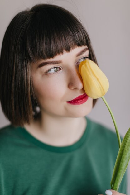 Close-upportret van betoverend donkerbruin vrouwelijk model in groene kledij ruikt tulp. zorgeloze vrouw met kort kapsel poseren met bloem.