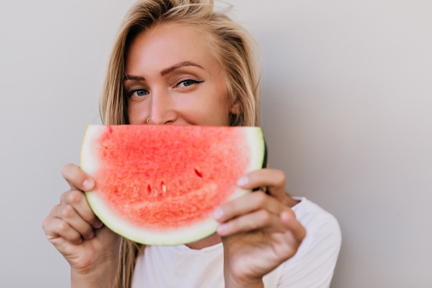 Close-upportret van blije blanke vrouw die fruit eet. Indoor foto van schattige blonde vrouw gek rond op lichte achtergrond.