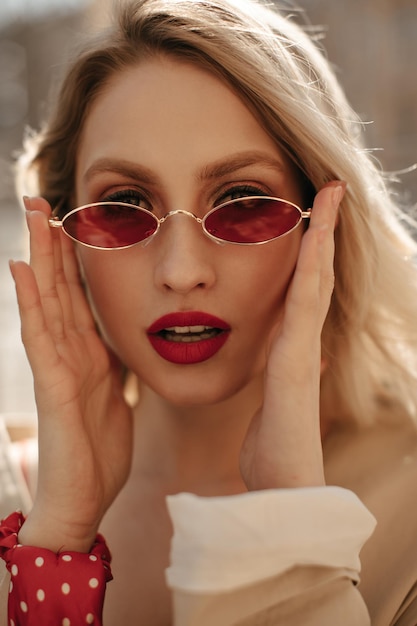 Close-upportret van blonde jonge vrouw met rode lippen die een heldere, stijlvolle, kleurrijke zonnebril opzet. elegante dame kijkt naar de camera buiten