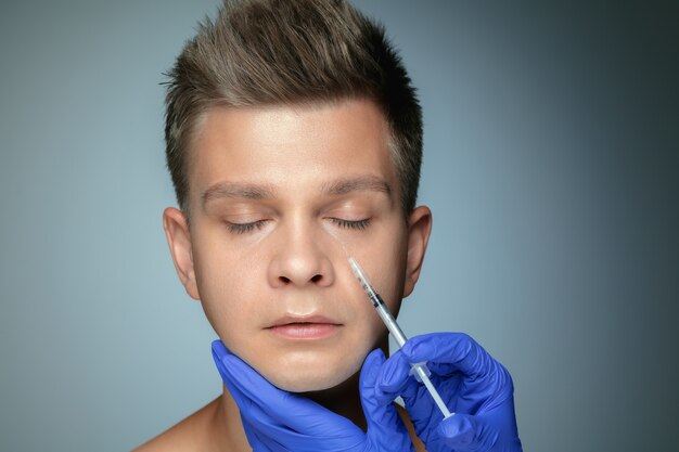 Close-upportret van de jonge mens die op grijze studiomuur wordt geïsoleerd bij het vullen van chirurgieprocedure