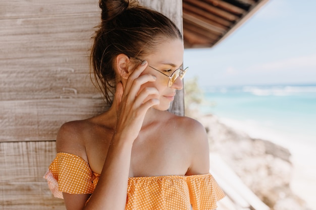 Gratis foto close-upportret van doordacht donkerbruin meisje dat zich op houten muur bevindt. buiten schot van verfijnde gelooide dame in oranje kleren wegkijken.