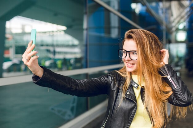 Close-upportret van een jong meisje dat zich buiten in luchthaven bevindt. Ze heeft lang haar, een zwarte jas en een bril. Ze maakt een selfie-portret.