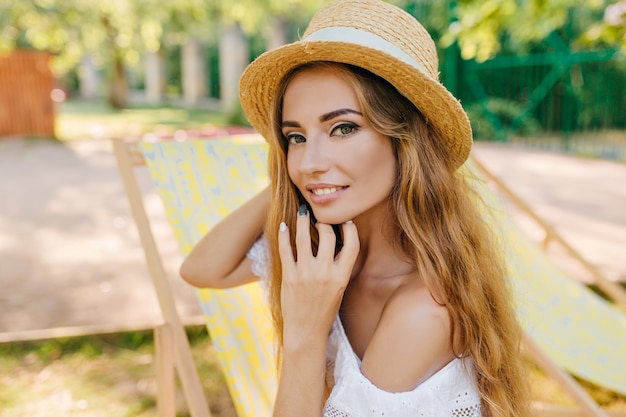 Close-upportret van geïnspireerd meisje met licht gebruinde huid die met haar lang glanzend haar speelt. Buiten foto van lachende jonge vrouw in vintage schipper en witte zomerjurk.