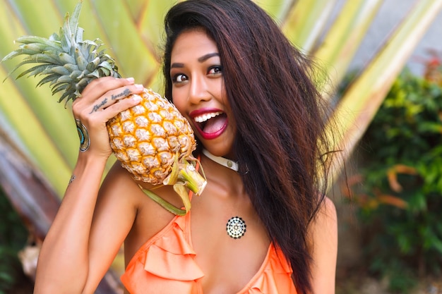 Close-upportret van gelooid Aziatisch meisje met vingertatoegering die bikini draagt. Vrij Latijns-jonge vrouw met ananas en lachen met palmboom op achtergrond.