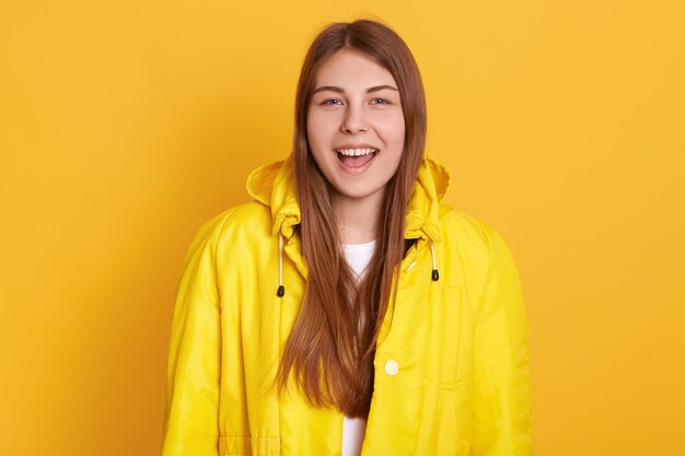 Close-upportret van gelukkig glimlachend studentenmeisje die jasje dragen, die gelukkig iets schreeuwen, status geïsoleerd over gele muur, die positieve emoties uitdrukken.