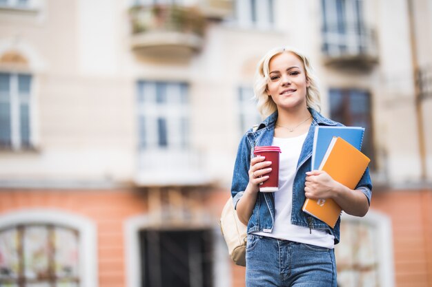 Close-upportret van gelukkige blonde studente met heel wat notitieboekjes gekleed in spijkerbroek