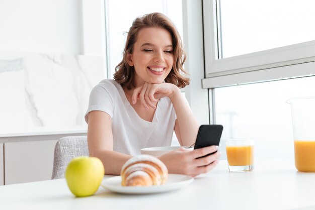 Close-upportret van gelukkige donkerbruine vrouw die mobiele telefoon met behulp van terwijl het hebben van ontbijt bij witte keuken