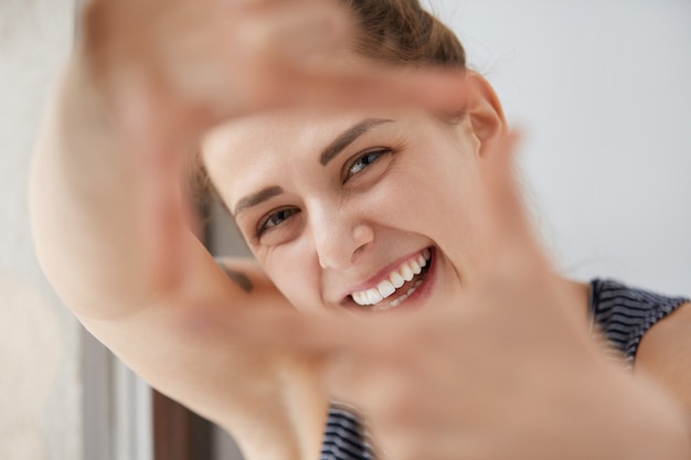 Close-upportret van glimlachend mooi glanzend Europees meisje door het kader van haar handen. Gelukkige vrouw met ogen dichtknijpen, plezier maken, fotograaf proberen te imiteren, speels zijn als een klein kind.