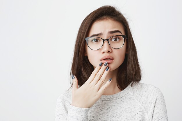 Close-upportret van jonge interessante vrouwelijke vrouw in glazen met verstoorde uitdrukking en opgeheven wenkbrauw, houdend vingers op kin